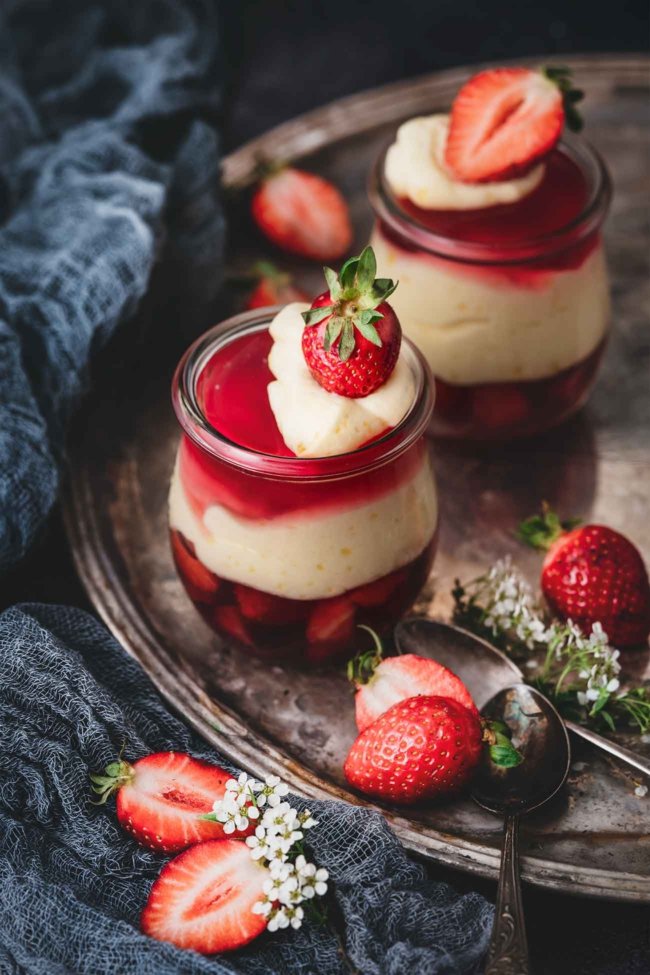 Götterspeise mit Orangen-Vanillepudding und Erdbeeren Jelly with orange custard and strawberries Jetzt wird es fruchtig im Glas: Himbeer, Erdbeer und Orange! Den Wunsch „ich möchte mal wieder Götterspeise essen“ habe ich erfüllt. Ich finde Götterspeise ehrlich gesagt ja ziemlich langweilig, aber in dieser Kombination finde ich sie richtig klasse. 4 Portionen 1 Beutel Himbeer Instant Götterspeisenpulver für 500 ml Wasser 1 Pck. Vanille-Puddingpulver zum Kochen 500 ml Orangensaft 100 g Zucker 300 ml Schlagsahne 250 g Erdbeeren, gewürfelt Puddingpulver mit dem Zucker und 6 EL Orangensaft verrühren Restlichen Saft aufkochen, angerührtes Puddingpulver zugeben und aufkochen lassen Eine Minute köcheln lassen, auskühlen lassen, dabei ab und zu umrühren 500 ml kochendes Wasser mit dem Instant Götterspeisenpulver verrühren und ca. die Hälfte der Flüssigkeit auf 4 Gläser aufteilen Erdbeerwürfel zugeben und kalt stellen 2/3 der Orangen-Vanillepudding auf die Gläser verteilen, danach die restliche Götterspeise darüber gießen, gefolgt von dem restlichen Pudding Alles im Kühlschrank fest werden lassen