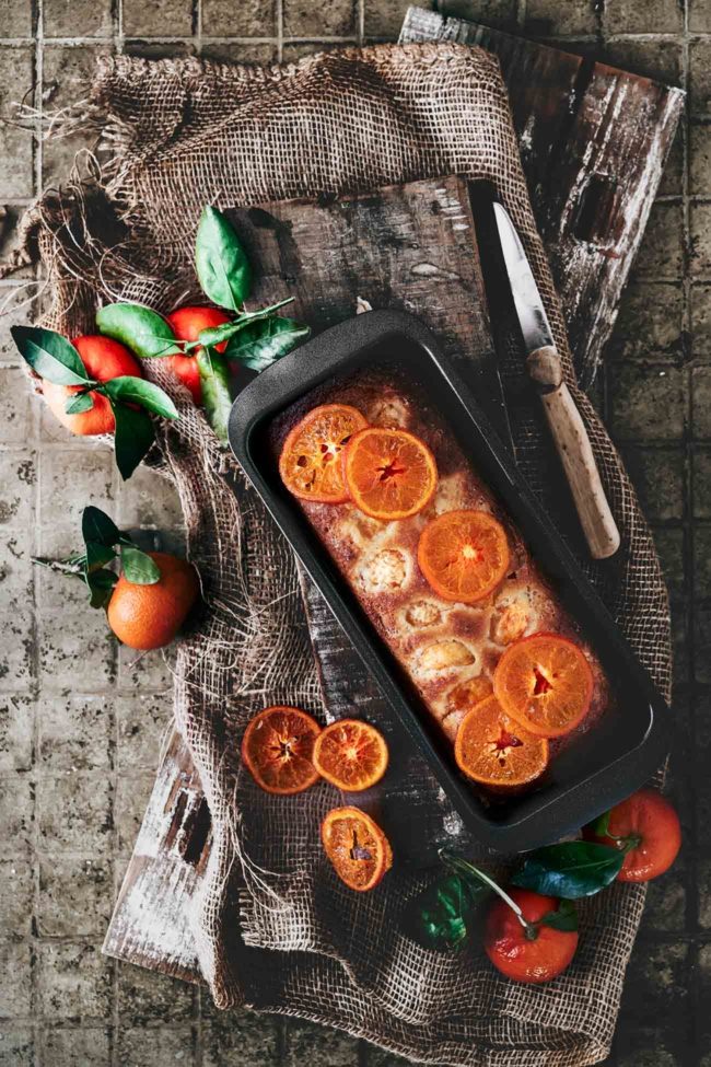 Mandarinen-Orangen Joghurt Kuchen Tangerine Orange Yoghurt Cake Dieser super softe Winter-Citrus Kuchen, mit einer Mischung aus Orangen und Mandarinen ist irgendwie warm und beruhigend. Ein toller Kuchen für das stürmische Wetter da draußen. 2 Orangen 4 Mandarinen 1 TL Vanilleextrakt 300 g Mehl 3 TL Backpulver 1 Prise Salz 500 g Zucker 150 g Joghurt (Vanille oder Natur) 5 Eier 150 g Olivenöl Orangen heiß abwaschen und die Schale fein abreiben, Saft auspressen und 200 ml Saft auffangen Saft und 200 ml Zucker in einen Topf geben und zum Kochen bringen, bis sich der Zucker vollständig aufgelöst hat Zwei Mandarinen mit Schale in feine Scheiben schneiden, dann zur Zuckermischung geben und 5 Minuten mit köcheln lassen Backofen auf 120 Grad Ober-Unterhitze vorheizen, Mandarinenscheiben entnehmen, abtropfen lassen und auf ein mit Backpapier ausgelegten Backblech legen und im Ofen 60 Minuten backen Backofen auf 180 Grad Ober-Unterhitze und eine Kastenform (Länge ca. 25 cm) mit Backpapier auslegen Orangenschale mit dem restlichen Zucker vermengen Eier mit Joghurt, Vanille und Olivenöl aufschlagen Mehl, Backpulver und Salz vermengen und nach und nach unterrühren Teig in die Form geben Restliche Mandarinen schälen und die einzelnen Mandarinenstükchen auf den Teig geben und ganz leicht andrücken Im Ofen 50-60 Minuten backen (Stäbchenprobe machen) und komplett in der Form auskühlen lassen Vor dem Servieren mit den kandierten Mandarinenscheiben und/oder dem restlichen Sirup beträufeln