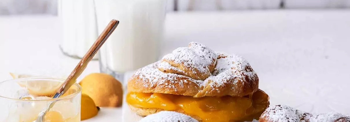 Crogels mit Lemon Curd Füllung Crogels with lemon curd filling 5 Zutaten und nur ein paar Handgriffe. Fertig ist die Mischung aus Croissant und Bagel, gefüllt mit Zitronencreme. Garantiert einfach! Einfach lecker. 6 Stück 2 Dosen frischer Croissantteig, Kühlregal für je 6 Croissants 1 Eigelb 2 EL Milch 200 g Lemon Curd Puderzucker zum Bestäuben Backofen auf 200 Grad Ober-Unterhitze vorheizen Teige aus der Verpackung nehmen, ausrollen und jeden Teig in 3 lange Streifen schneiden Teigstücke mit der langen Seite zusammenklappen und auf ca. 30 cm ausrollen/ziehen Jeden Teigstrang zu einer 20 cm langen Kordel drehen, dann zu einem Knoten formen 1,5 Liter Salzwasser zum Kochen bringen und jeden Teigring von jeder Seite 10 Sekunden garen, herausnehmen, abtropfen lassen und auf ein Backblech legen Eigelb mit Milch verquirlen und die Teigknoten damit einpinseln Im Ofen 15 Minuten goldbraun backen, abkühlen lassen, dann durchschneiden und die untere Hälfte mit Lemon Curd bestreichen Obere Hälfte daraufsetzen und mit Puderzucker bestäuben