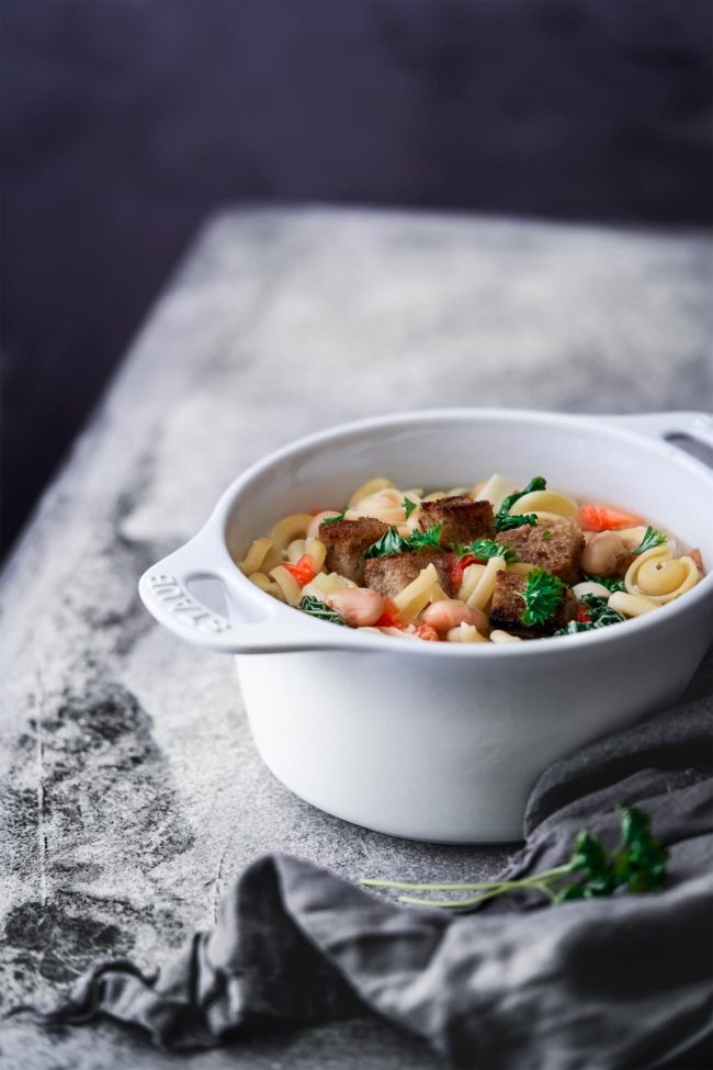 Minestrone aus dem Ofen Oven minestrone Diese Minestrone aus dem Ofen ist die perfekte Schüssel zum Aufwärmen an kalten Herbst-und Wintertagen. Jede Portion wird getoppt mit knusprigen Parmesan-Croutons und frischer Petersilie. Diese Schüssel Minestrone ist eine gemütliche Mahlzeit, bei der man sich einfach wohlfühlen muss. 1,5 Liter Gemüsebrühe 1 Zwiebel, gewürfelt 2 Möhren, gewürfelt ¼ Knollensellerie, gewürfelt 300 g Wirsing, in Streifen geschnitten 1 Lauch, halbiert und in Ringe geschnitten 2 Knoblauchzehen, gehackt 3 Tomaten, gewürfelt 400 g weiße Bohnen, aus der Dose 100 g Pasta ½ Bund Petersilie 200 g Bauernbrot, gewürfelt 25 g Parmesan, gerieben 1 EL Butter Backofen auf 200 Grad Ober-Unterhitze vorheizen. Gemüsebrühe in einen ofenfesten Topf zum Kochen bringen. Zwiebel. Möhren, Sellerie, Wirsing, Lauch, Knoblauch, Tomaten, Bohnen und Pasta in den Topf geben und alles 50 Minuten mit geschlossenem Deckel garen. In der Zwischen die Butter in einer Pfanne erhitzen und die Brotwürfel anbraten, Parmesan darüberstreuen. Suppe mit Croutons und Petersilie servieren.