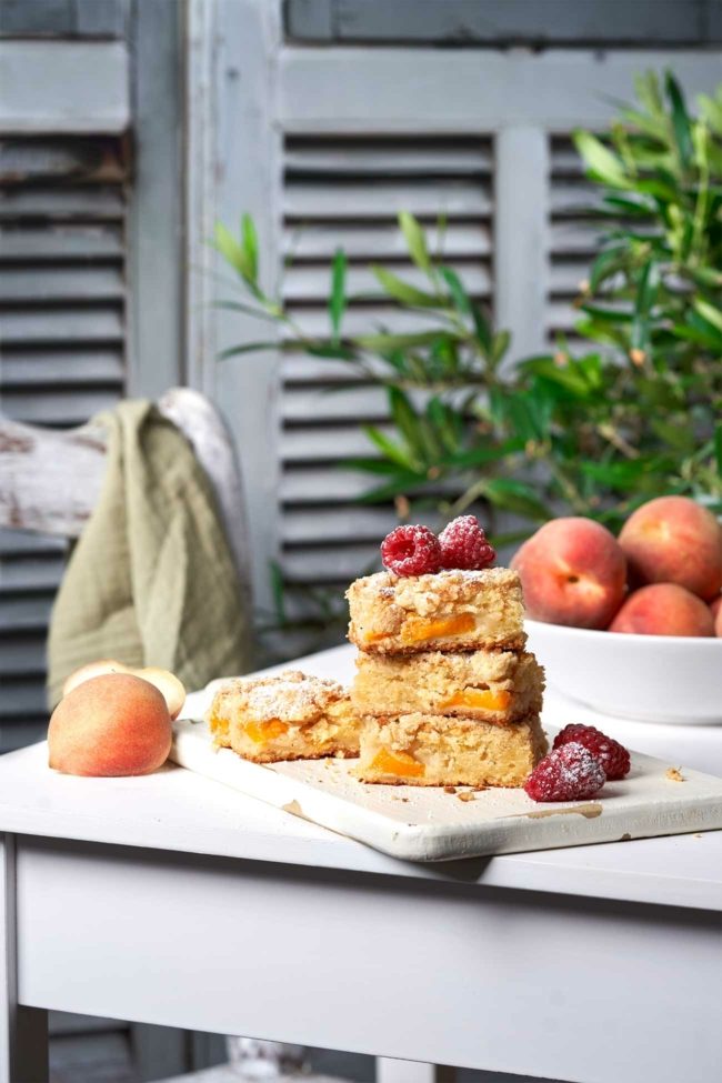 Pfirsichkuchen angerichtet auf einem weißen Tisch mit Pfirsichen im Hintergrund